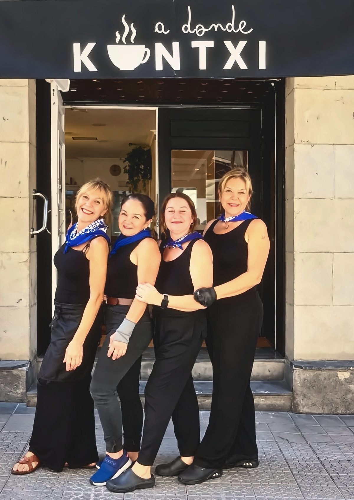 Las cuatro del equipo de A donde Kontxi, de cuerpo entero, sonrientes y con pañuelos azules, en la puerta de la cafetería bajo el letrero